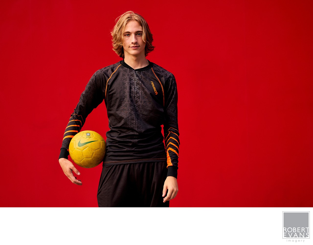 A young man dressed in black poses with a soccer ball beside a red wall.