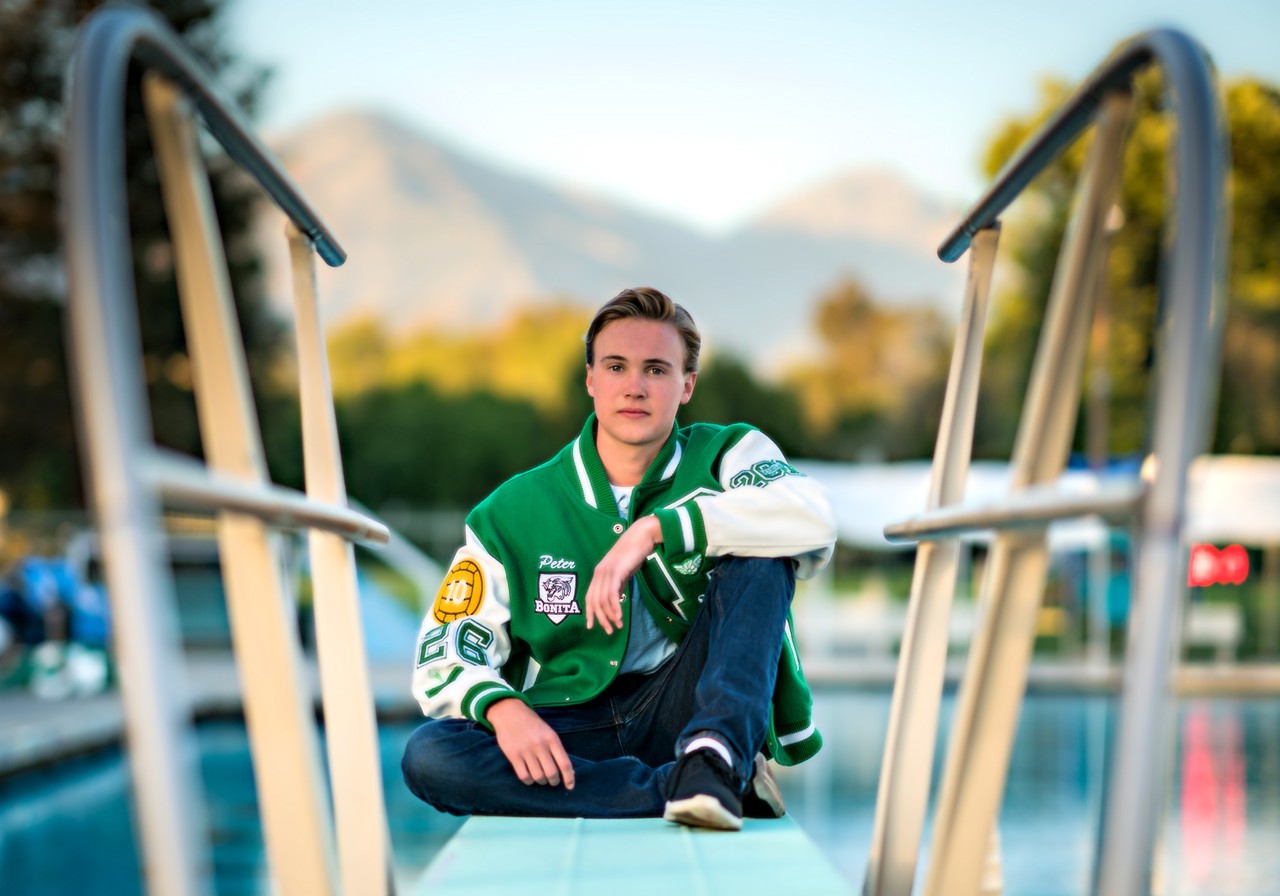A young man in a green letter jacket sits between swimming pool handrails.