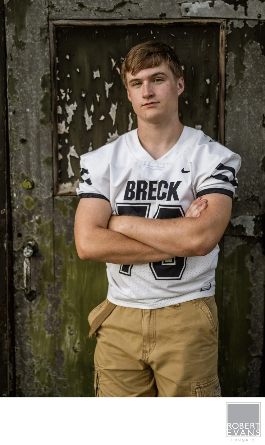 A young man in a Breck football jersey stands with folded arms at a weathered doorway.