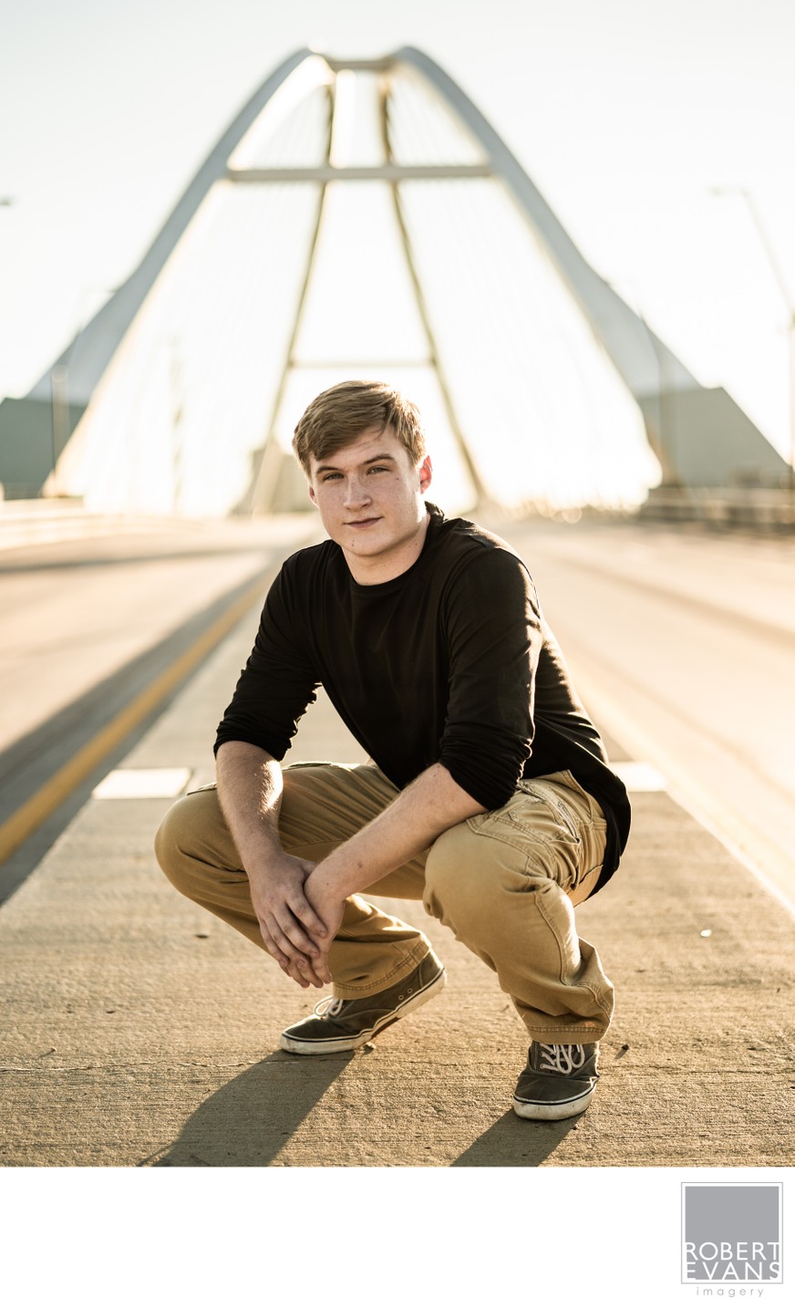 A young man in a black sweater crouches on a bridge beneath its cables.