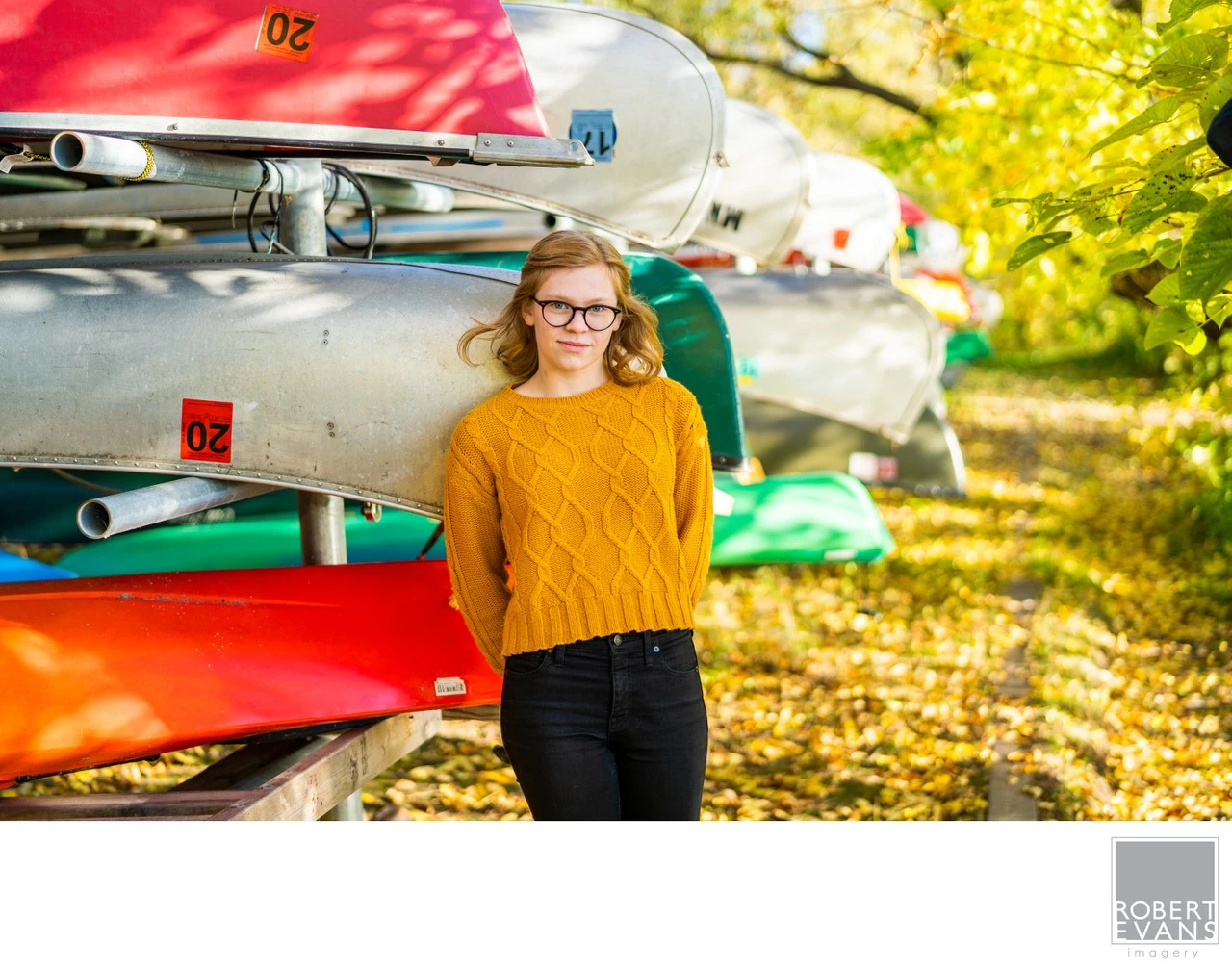 A young woman in glasses and a mustard sweater stands beside stacked colorful kayaks.