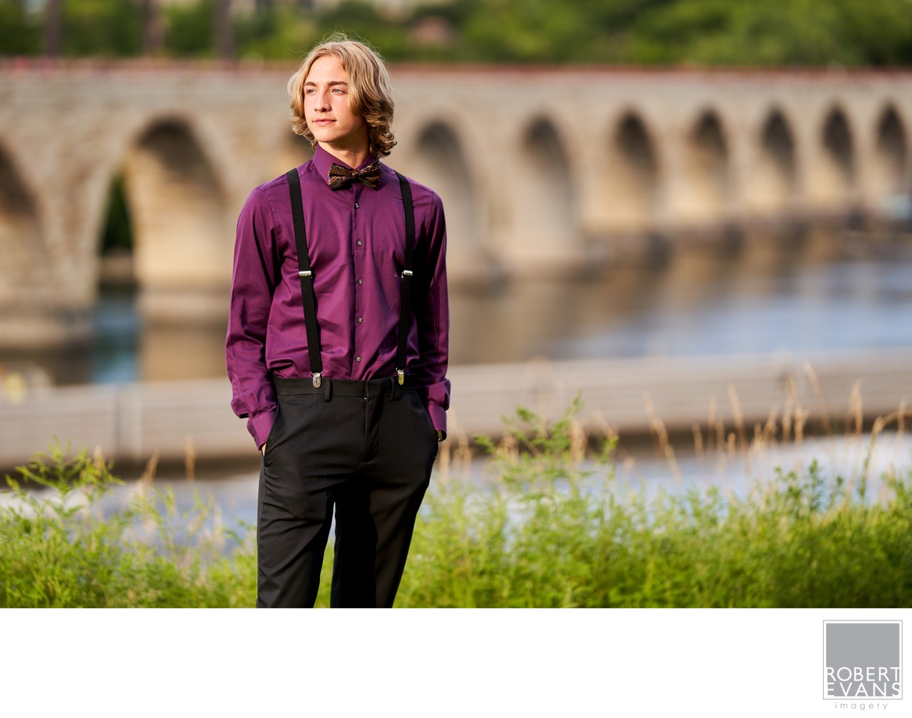 A young man in a purple shirt stands beside the river with the Stone Arch Bridge behind him.
