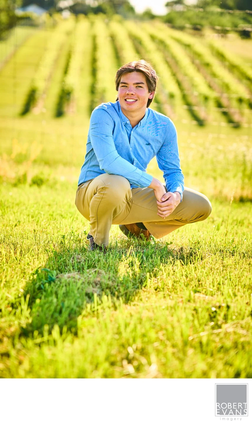 A young man in a blue shirt crouches in grass with vineyard rows behind him.