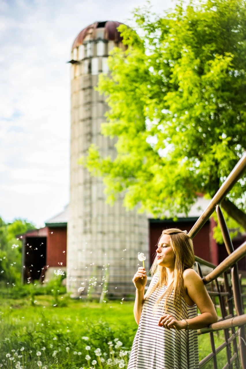 A young woman blows a dandelion beside a rustic fence and farm silo.
