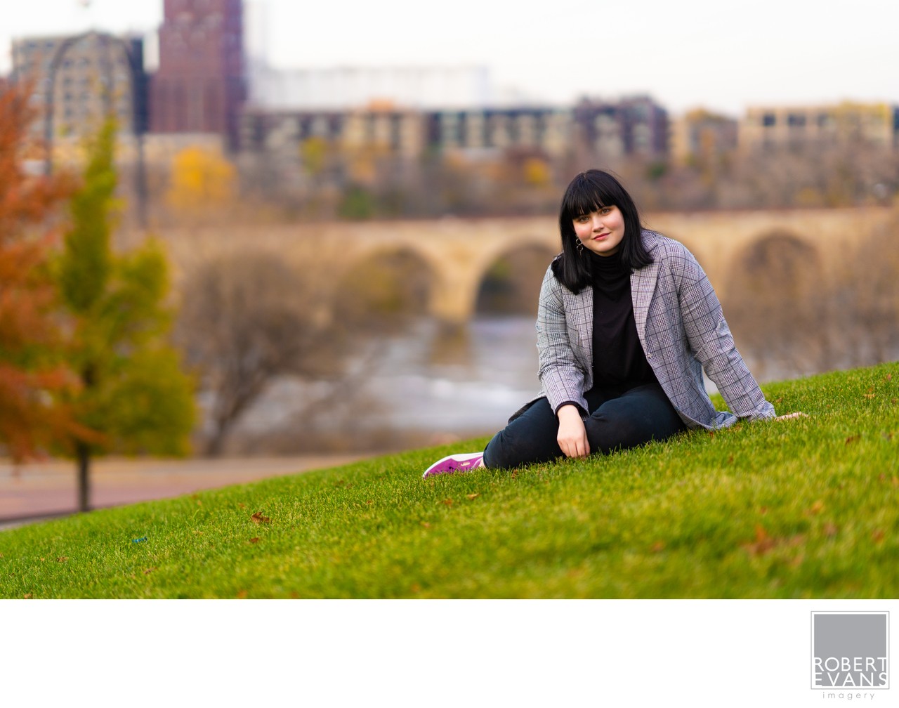 A young woman in a gray coat sits on a lawn overlooking the Stone Arch Bridge.