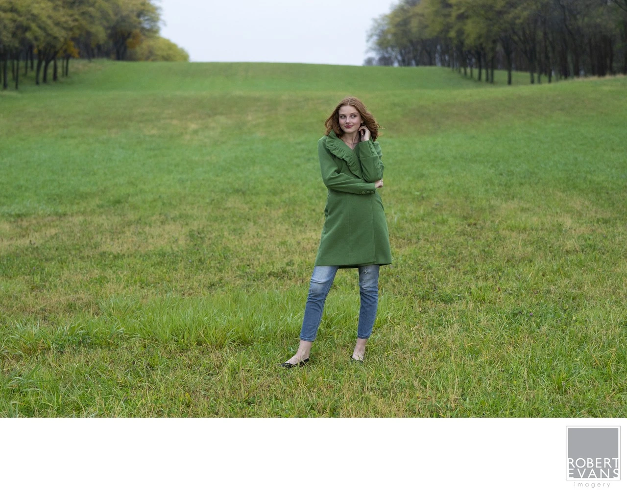 A young woman in a green coat stands in a broad grassy field.