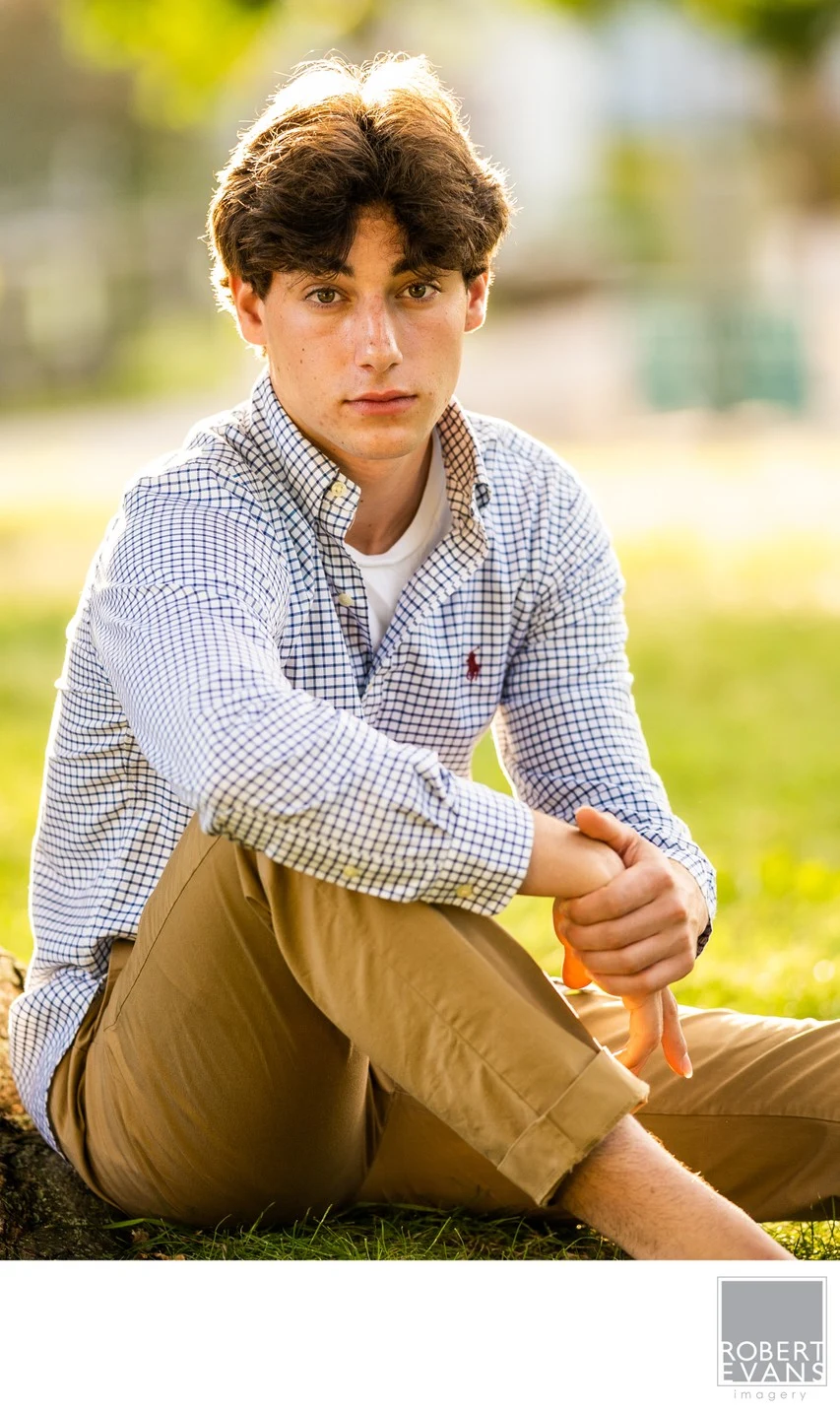 A young man in a patterned shirt and tan trousers sits outdoors in soft backlight.