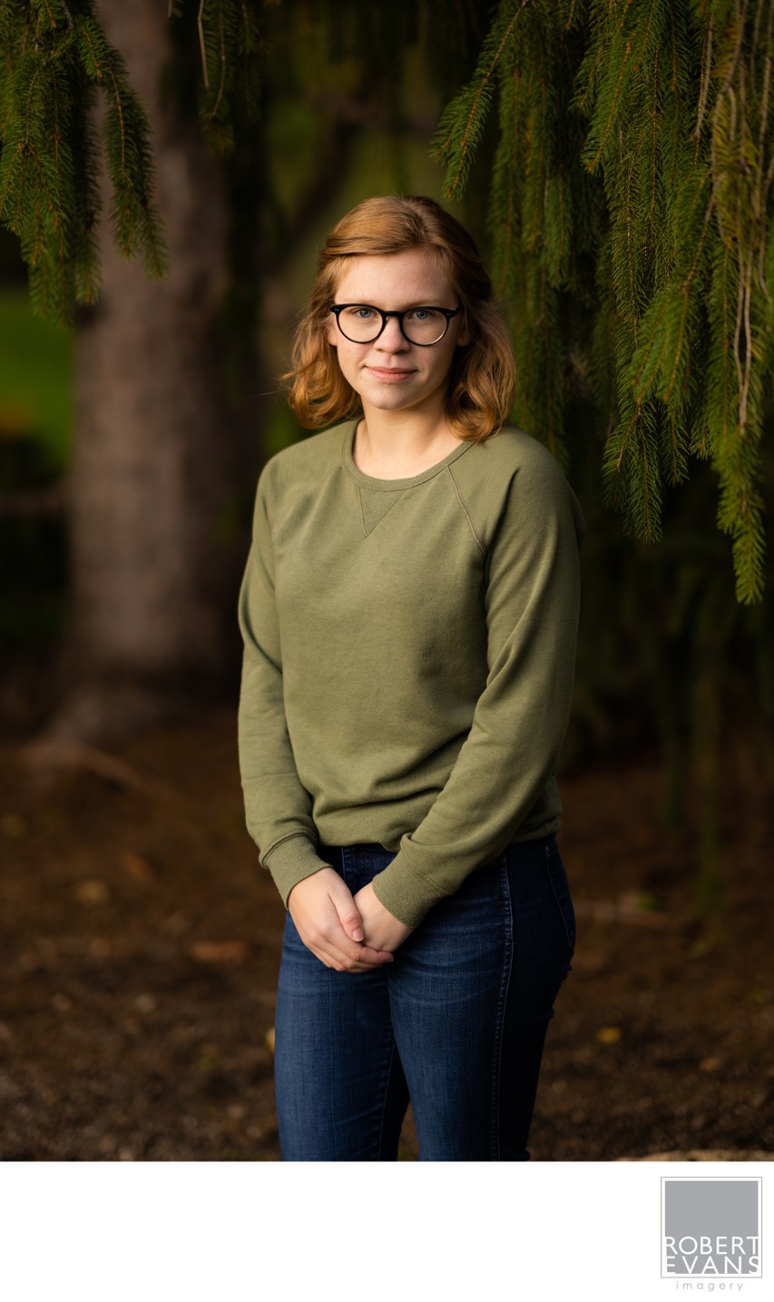A young woman wearing glasses and a green top stands beside a mossy tree trunk.