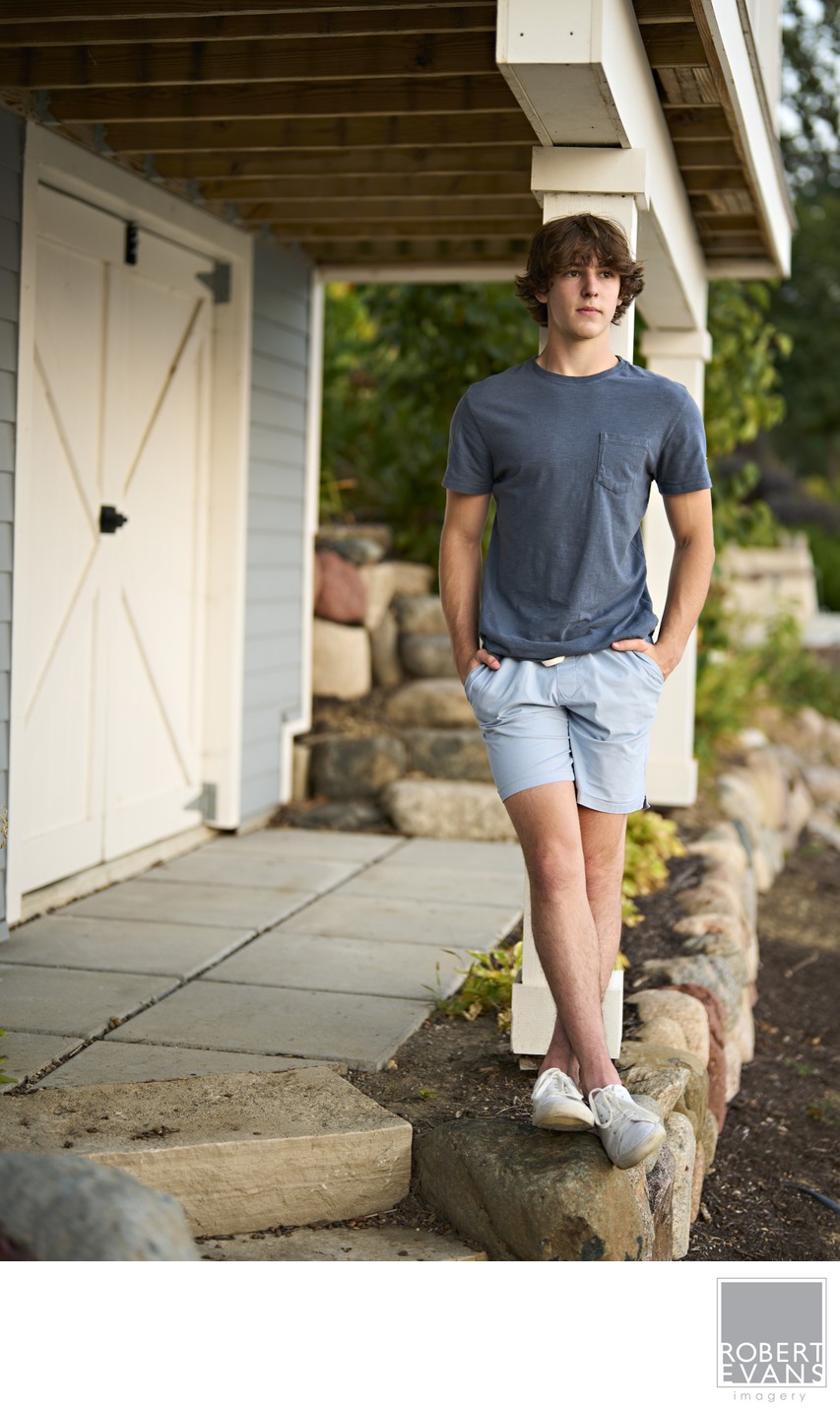 A young man in a gray shirt and shorts sits on a low stone wall beside a building.