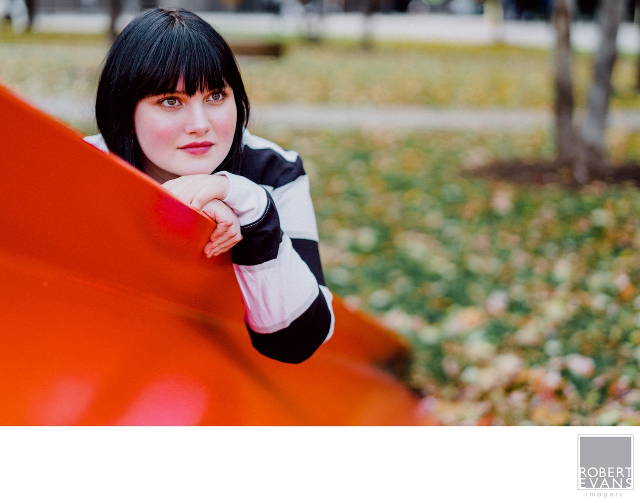 A young woman rests her hands on a red metal sculpture in an autumn setting.