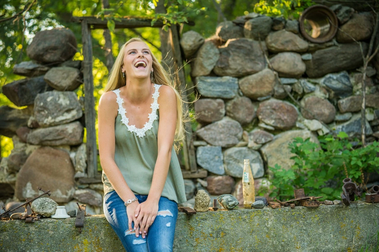 A young woman laughs while seated beside a rustic stone wall.