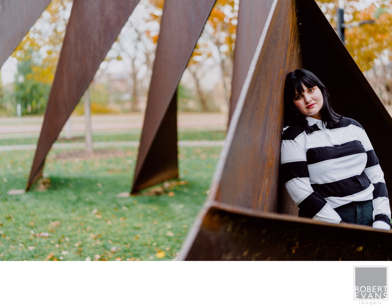 A young woman in a striped sweater poses within a large angular steel sculpture.
