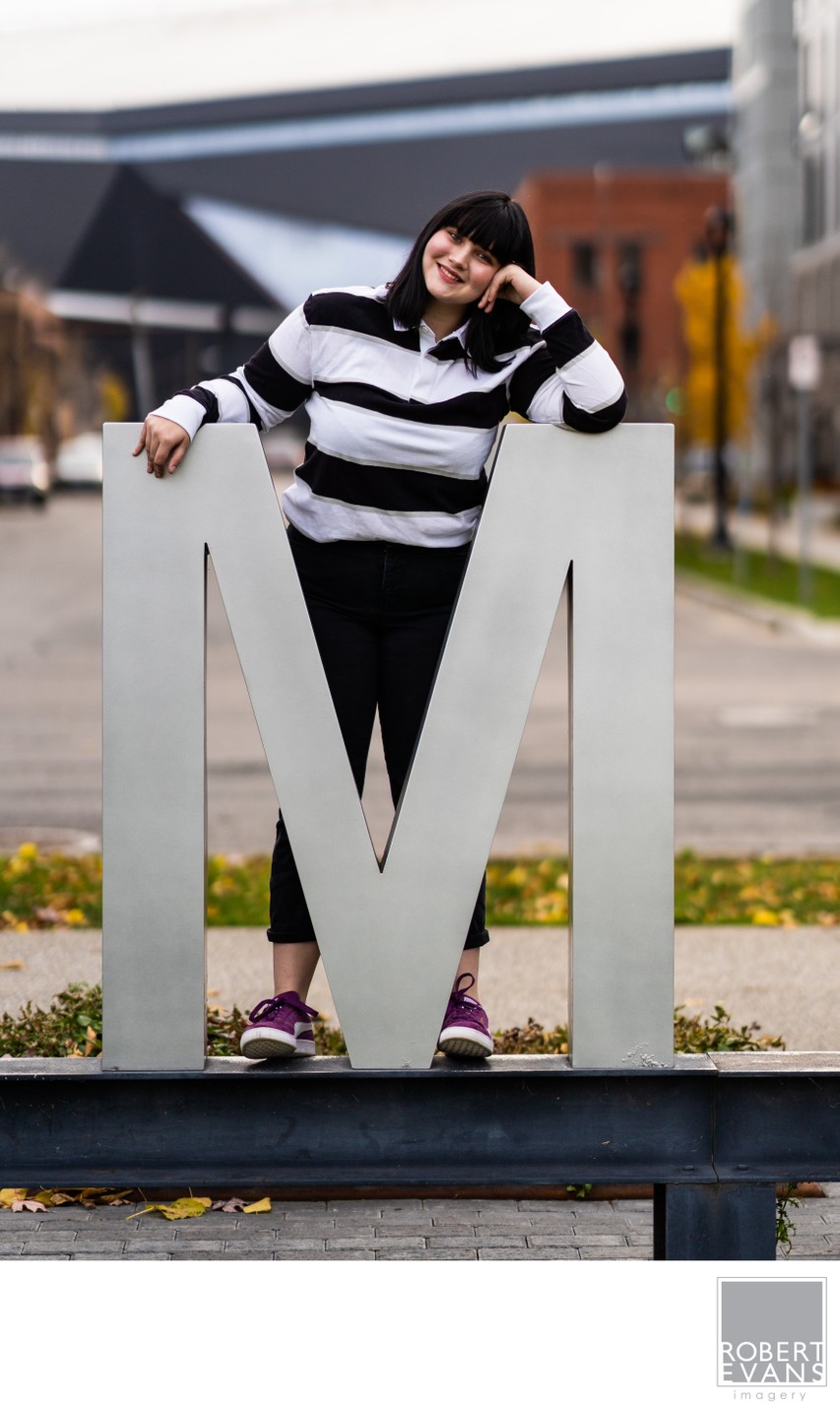 A young woman in a striped sweater leans against a large letter M sculpture.