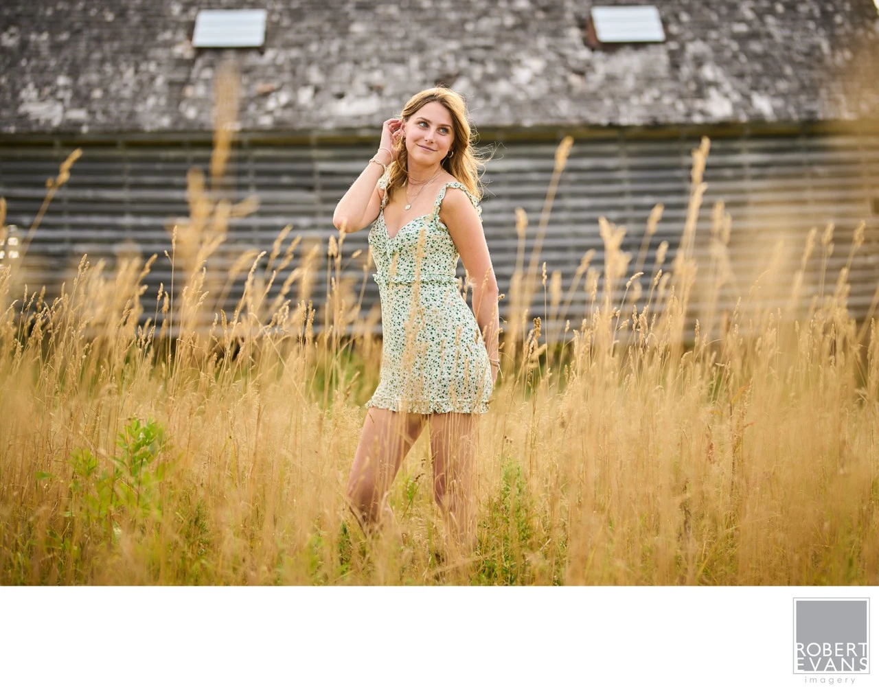 A young woman in a floral dress stands among grasses beside a weathered barn.