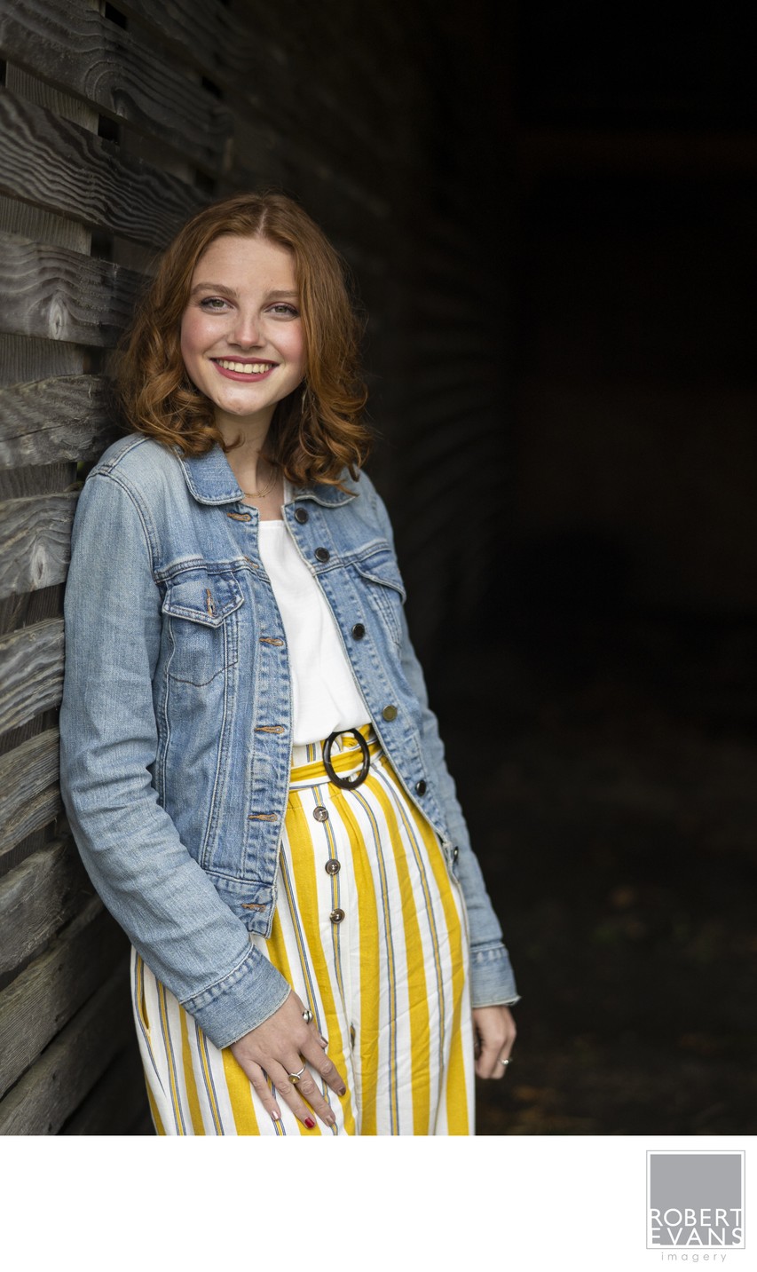 A smiling young woman wears a denim jacket and striped trousers in a dark wooden doorway.