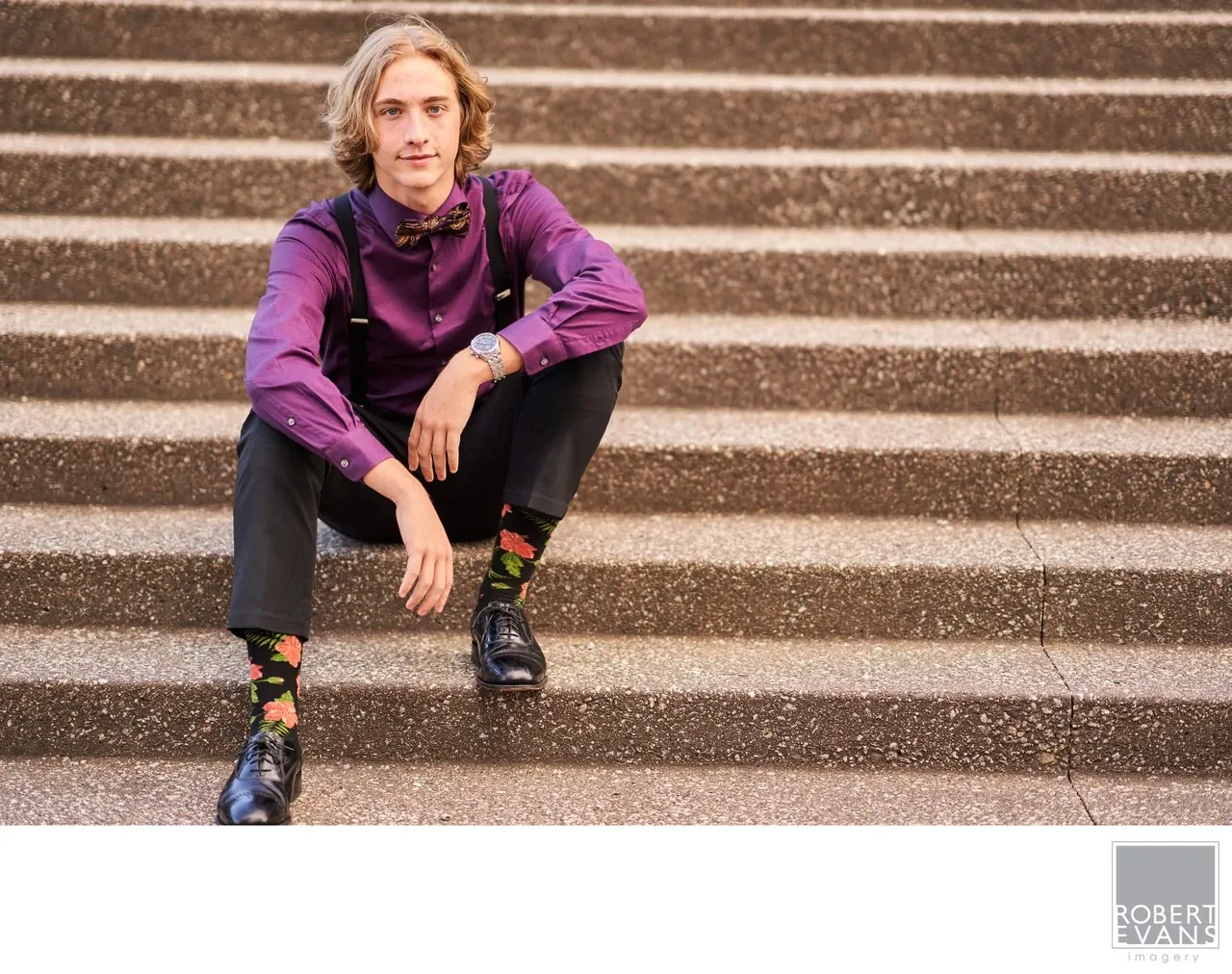 A young man in a purple shirt and suspenders sits on broad stone steps.