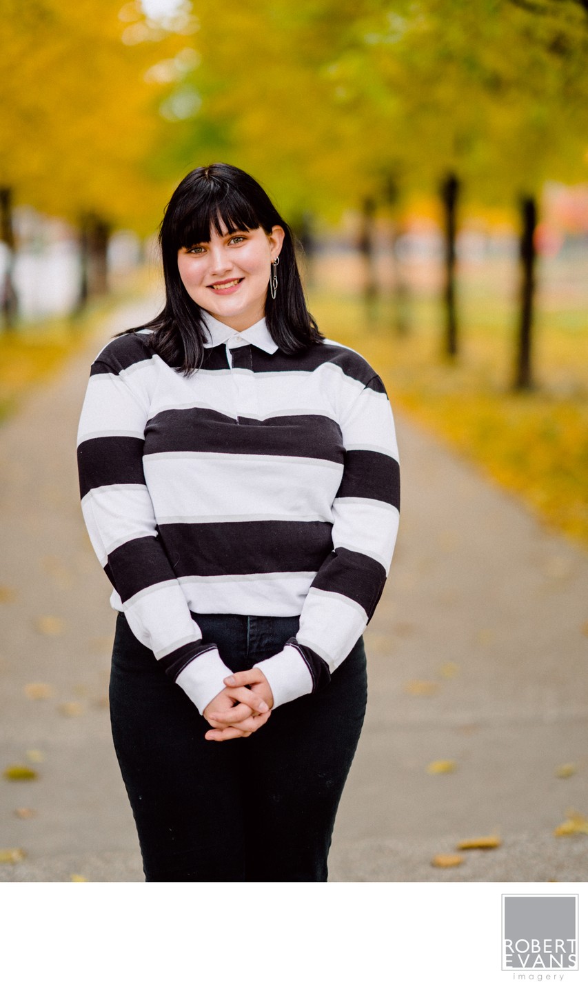 A young woman in a black-and-white striped sweater stands on an autumn path.