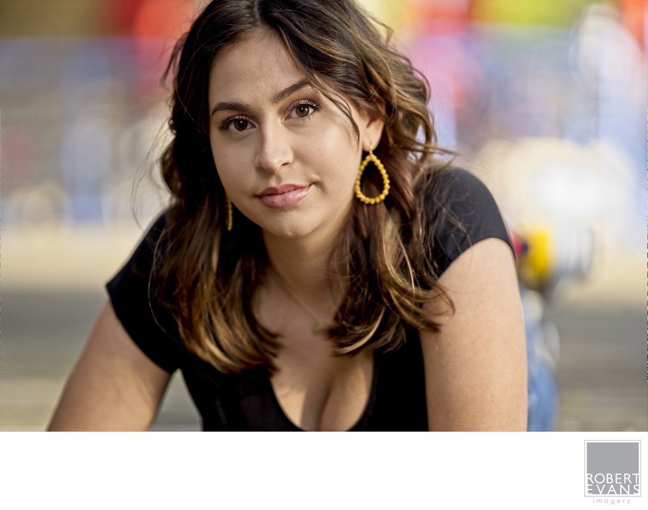 A young woman wearing gold hoop earrings leans toward the camera against a softly blurred background.