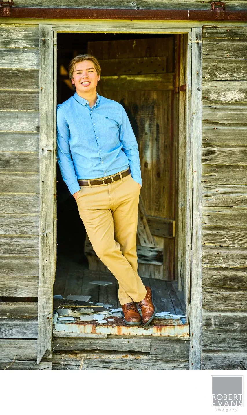 A young man in a blue shirt and tan trousers leans in a weathered wooden doorway.