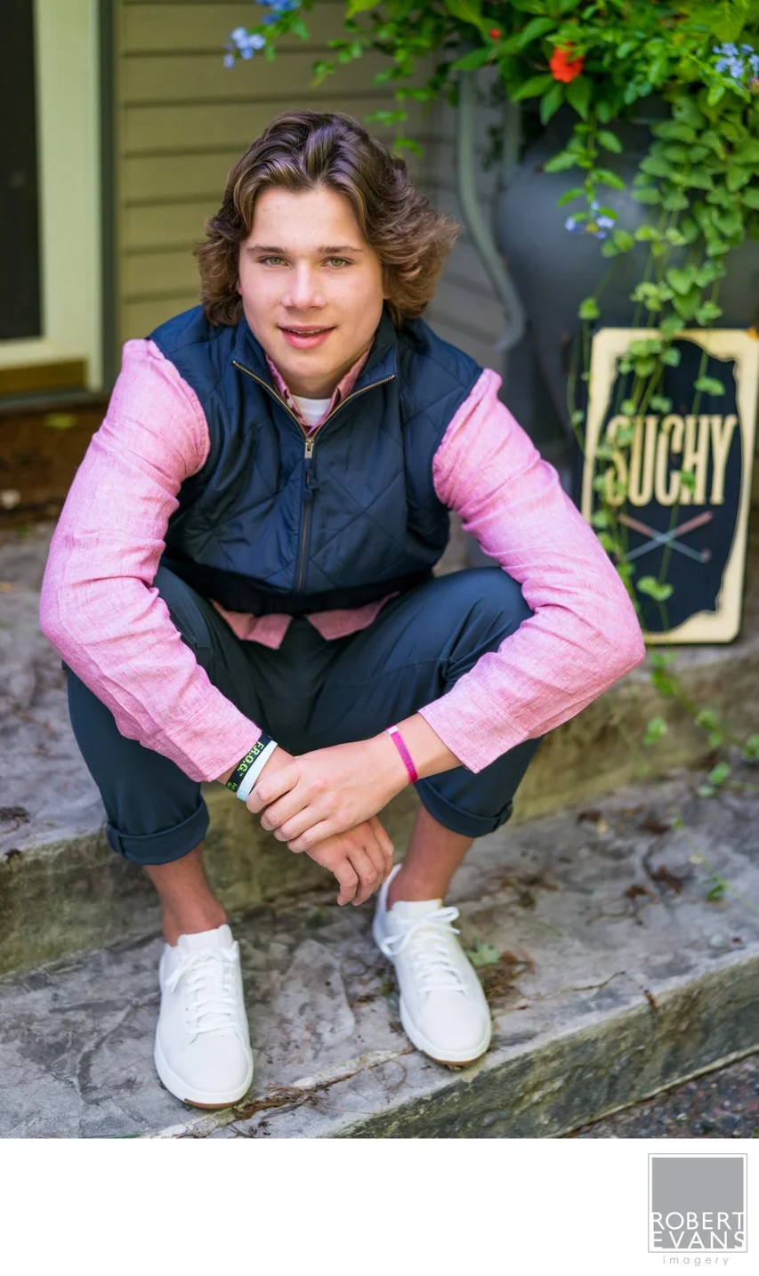 A young man in a pink shirt and dark vest crouches outside a storefront.