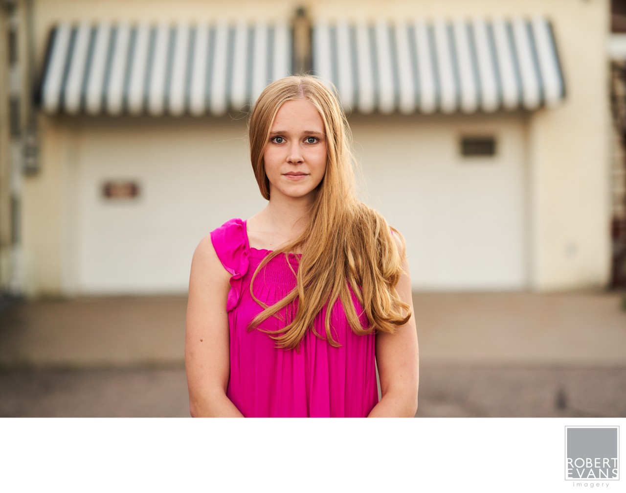A young woman in a pink dress stands in front of a building with a striped awning.
