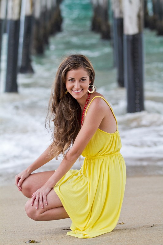 A young woman in a yellow dress crouches on sand beneath pier supports.