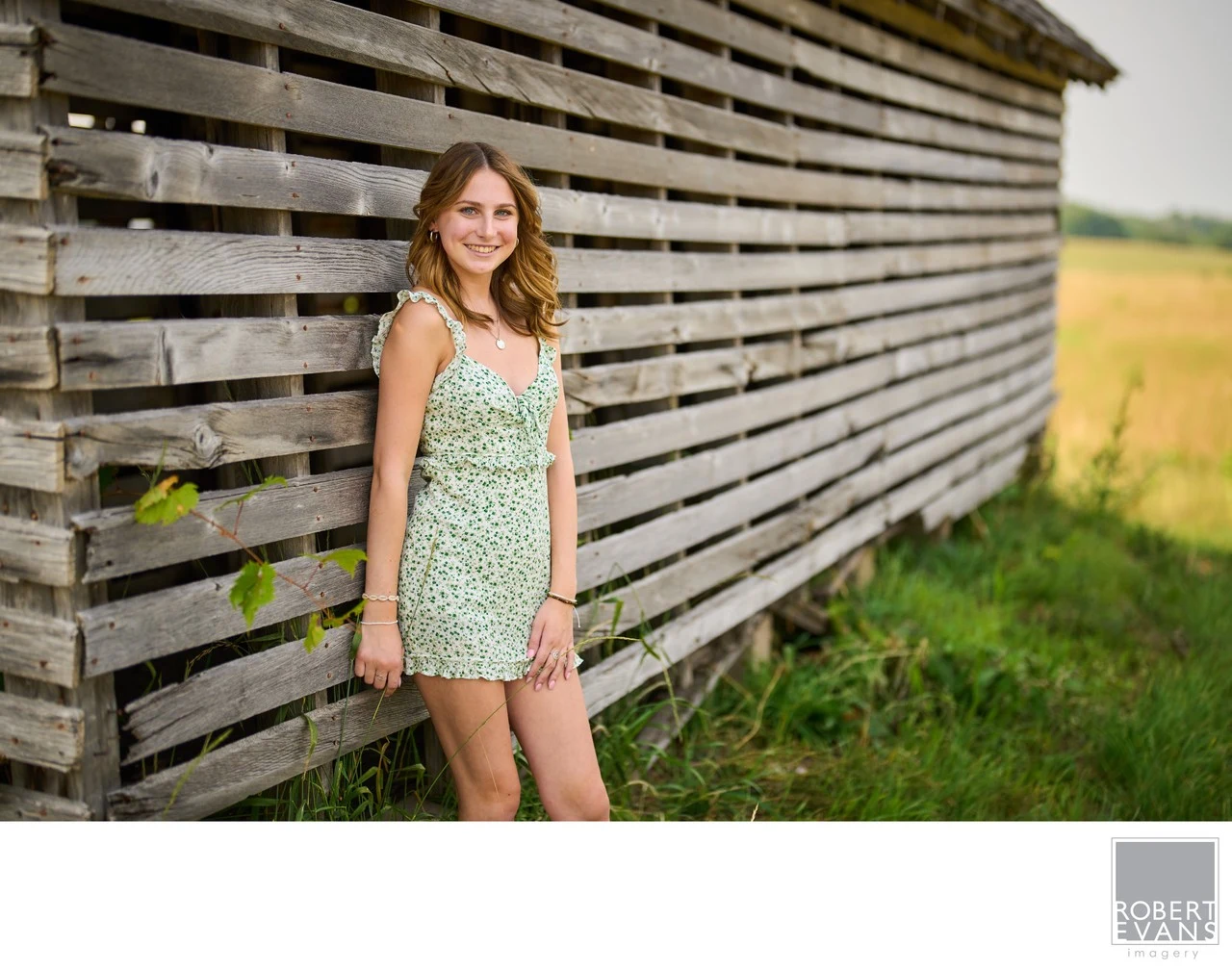 A young woman in a floral dress stands beside a wall of weathered wooden slats.