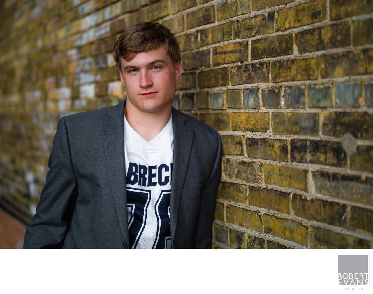 A young man wears a blazer over a Breck football jersey beside a brick wall.