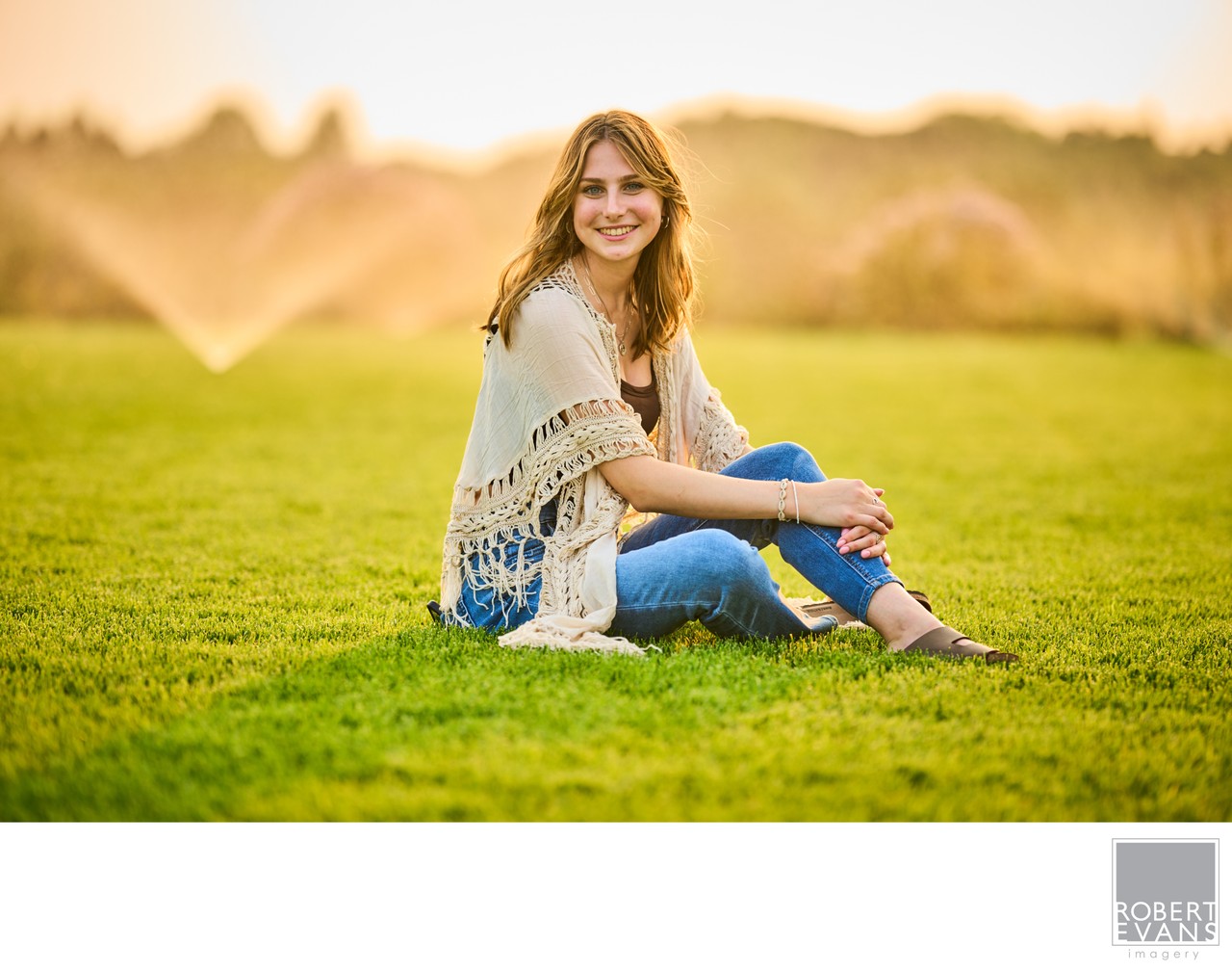 A young woman in jeans and a light shawl sits on grass in warm evening light.
