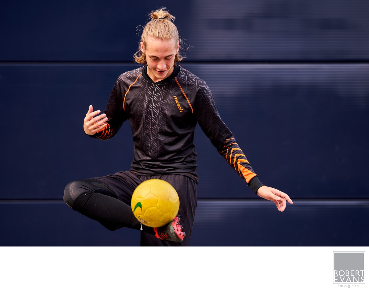 A young soccer player balances on one leg while controlling a yellow ball against a blue wall.