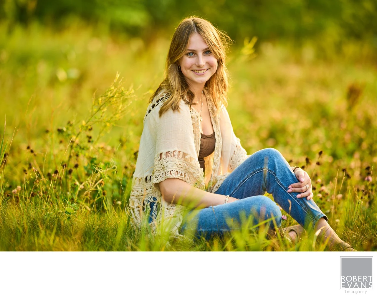 A young woman in jeans and a light shawl sits among tall green grasses.