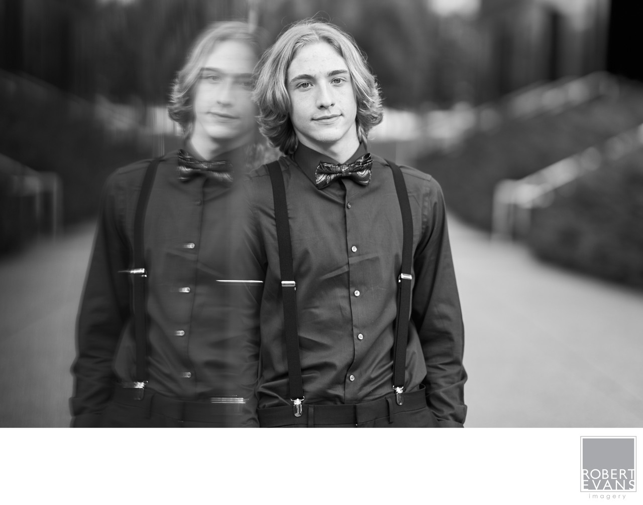 A young man in a bow tie stands beside a reflective window in black and white.