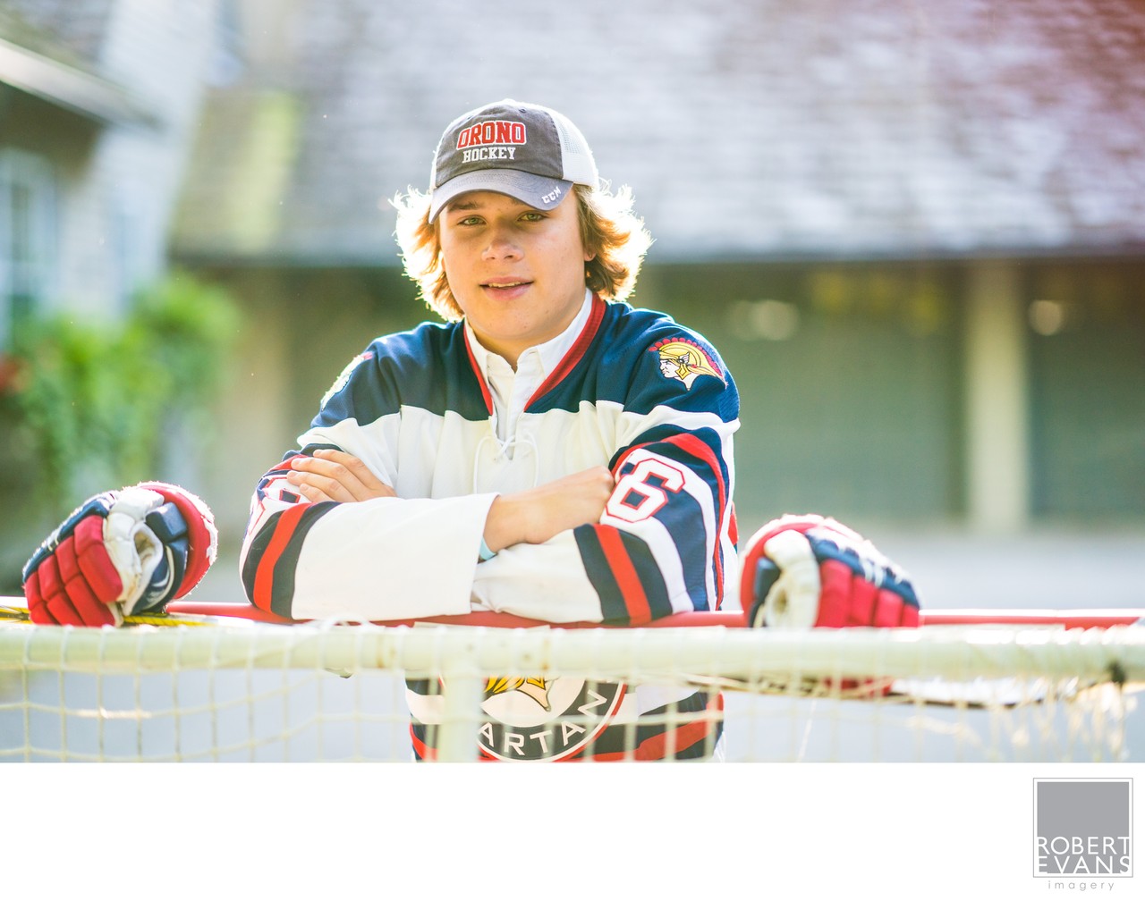 A young man wearing a hockey jersey and cap rests his gloves on a goal frame.