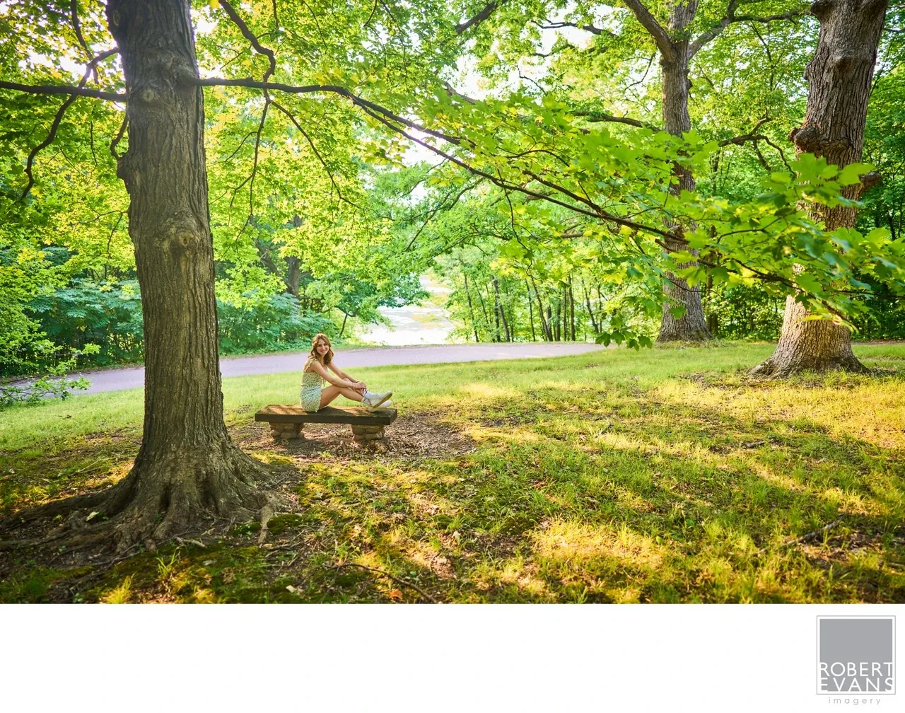 A young woman sits on a bench beneath broad leafy trees beside a lake.