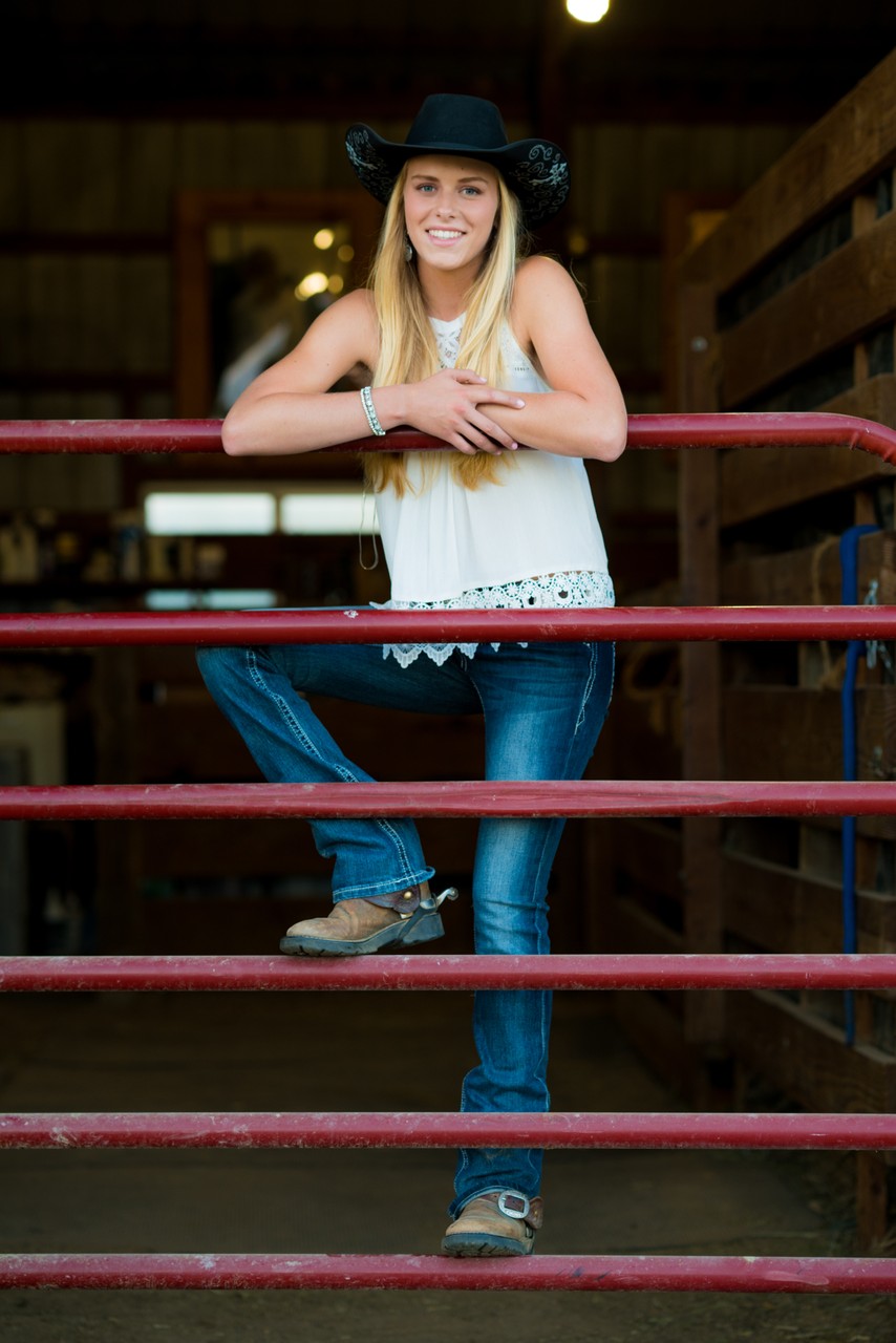 A young woman in a cowboy hat leans on a red gate inside a stable.