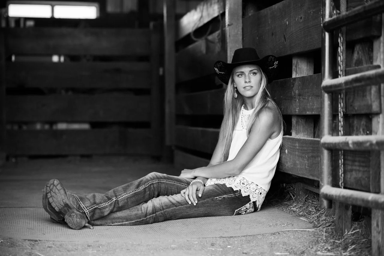 A young woman in a cowboy hat sits on the floor inside a wooden stable in black and white.