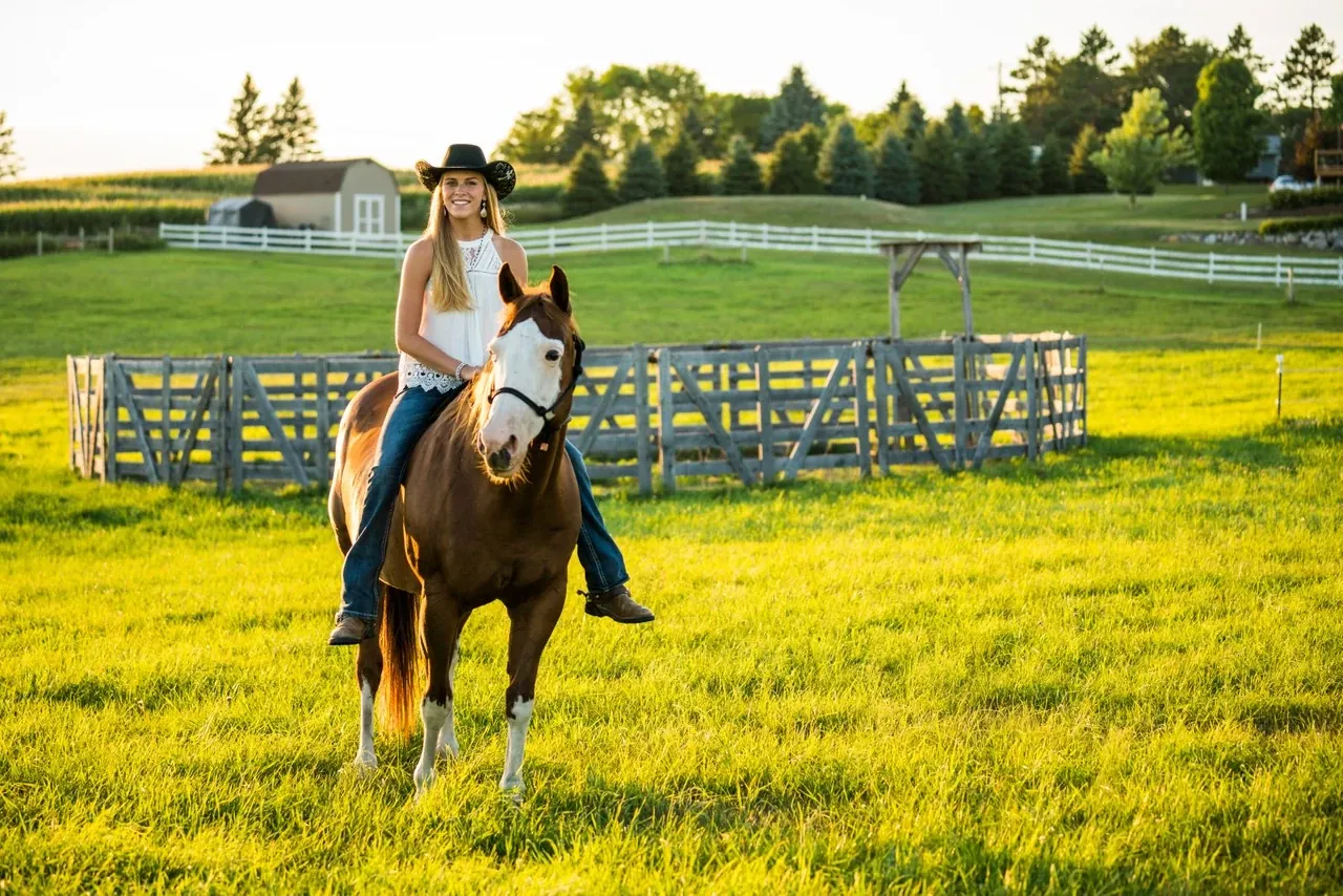 A young woman in a cowboy hat rides a horse through a sunlit green pasture.