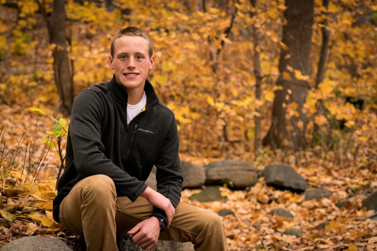 A young man in a black sweater sits among golden autumn leaves beside a stream.