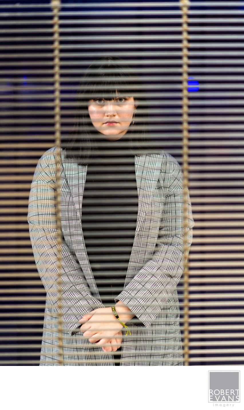 A young woman in a gray coat stands behind horizontal window blinds.