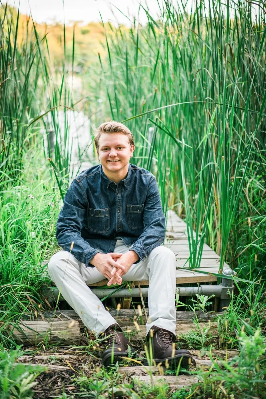 A young man sits on a wooden dock surrounded by tall green reeds.