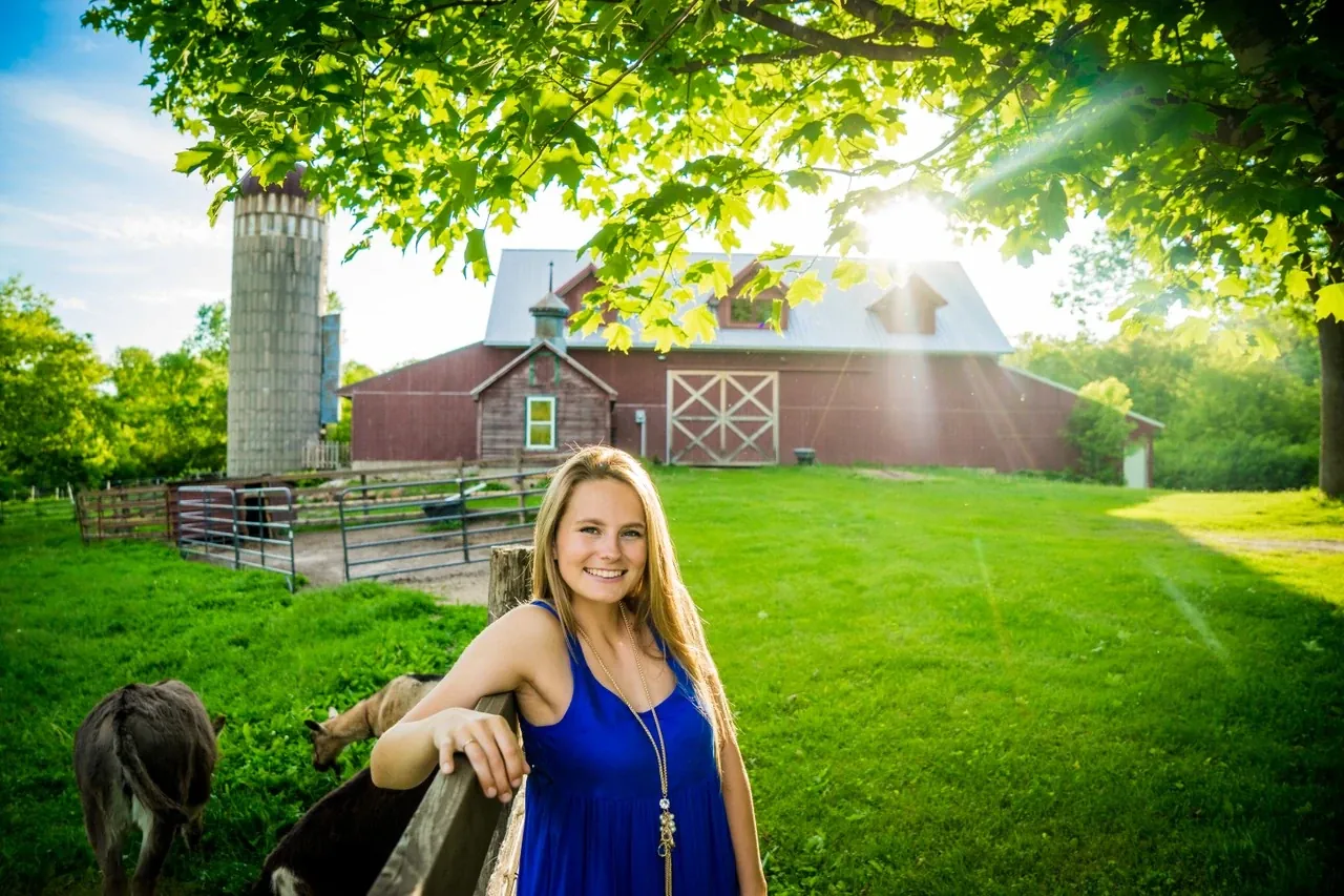 A young woman in a blue dress leans on a farm fence with a red barn behind her.
