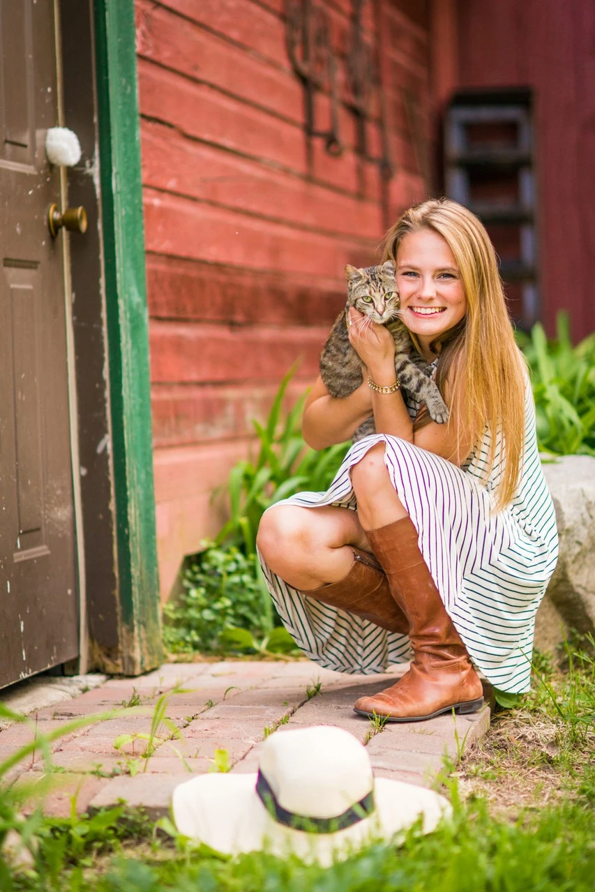 A young woman crouches beside a red barn holding a kitten, with a hat on the ground.