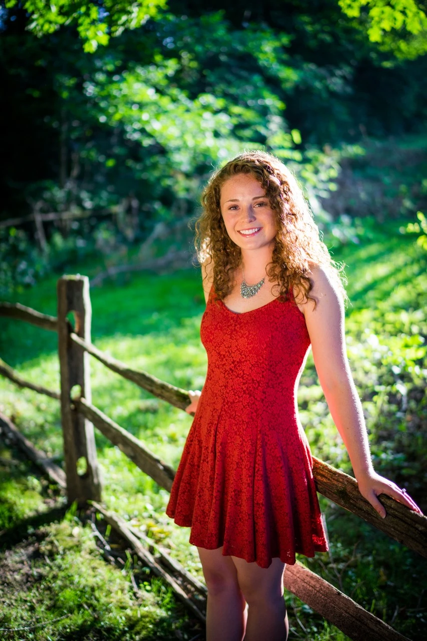 A young woman in a red dress leans against a rustic fence beneath green trees.