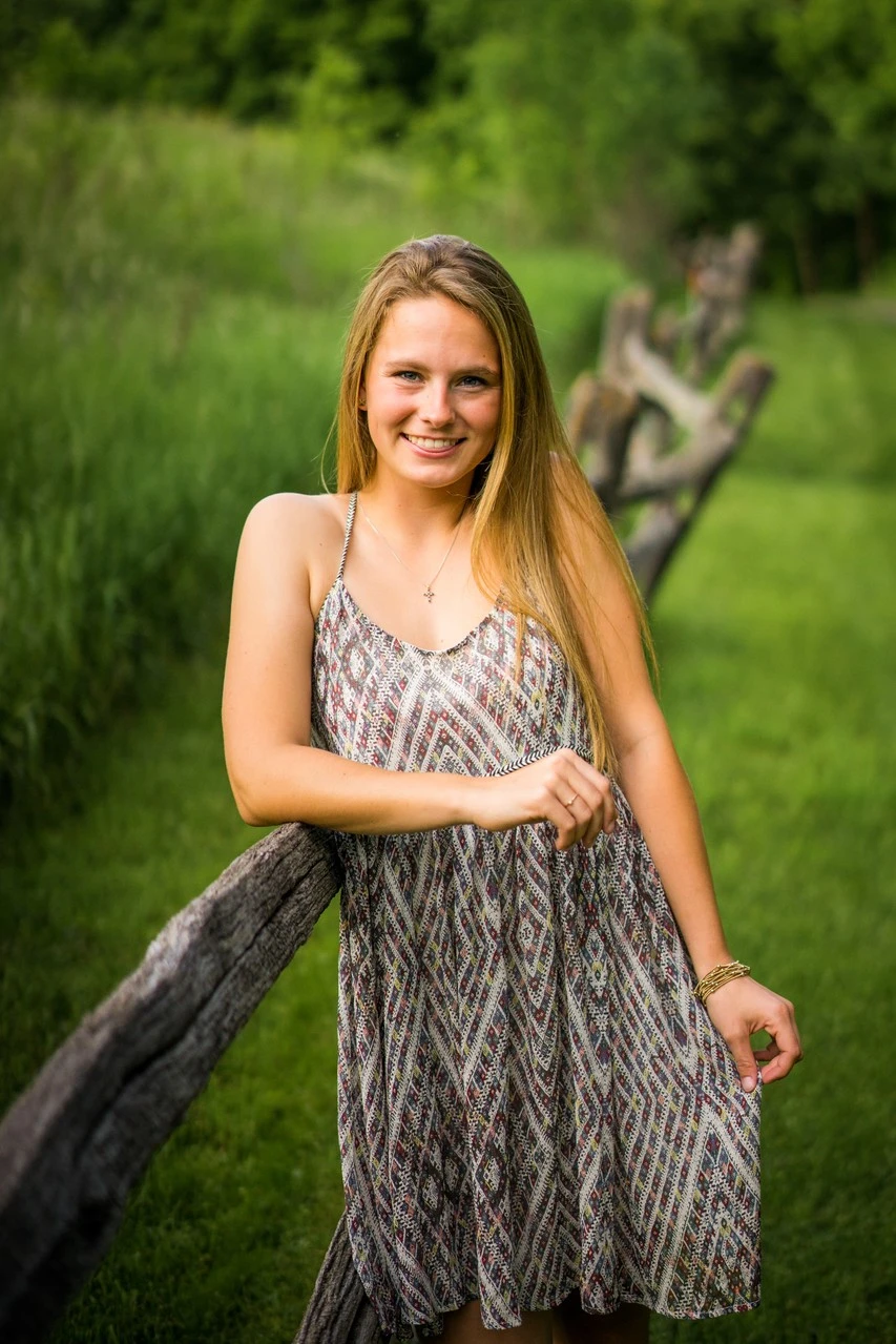 A young woman in a patterned dress rests an arm on a weathered wooden fence.