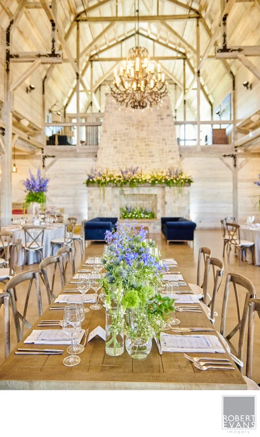 A long reception table with flowers and glassware beneath chandeliers in a timber-framed room.