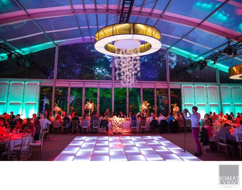 A reception tent with a central chandelier, dance floor and colorful lighting.