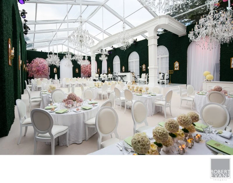 A reception room with white chairs, pink flowers and greenery beneath a glass roof.
