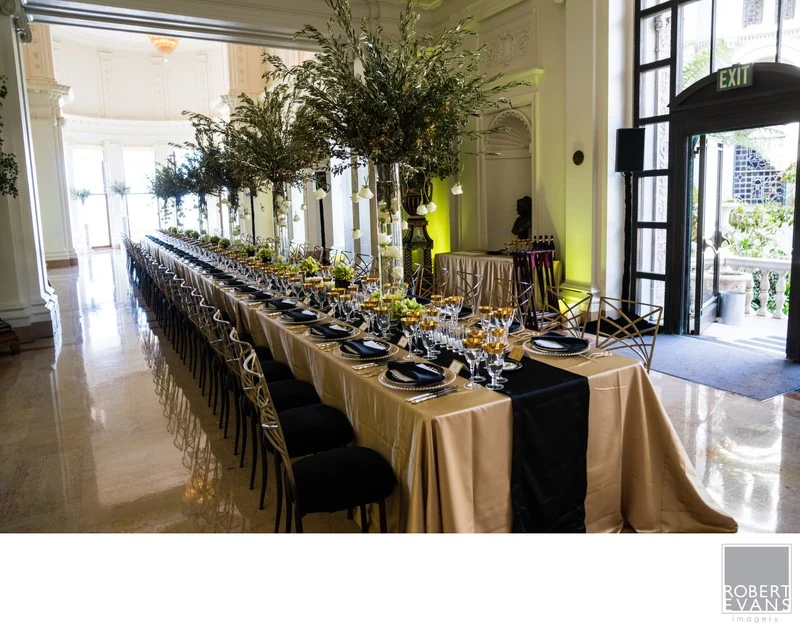 A long reception table with tall green centerpieces, glassware and dark chairs in a bright room.