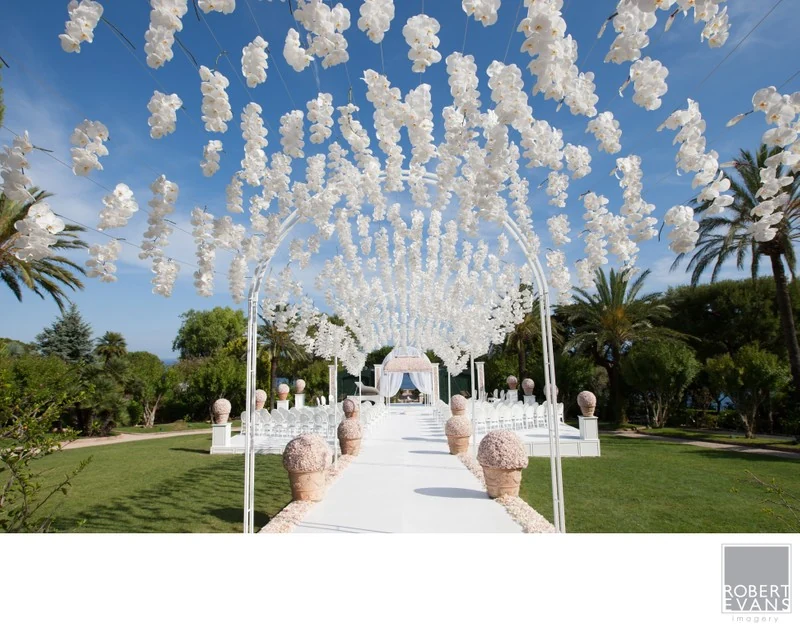 White flowers suspended above an outdoor ceremony aisle lined with white chairs.