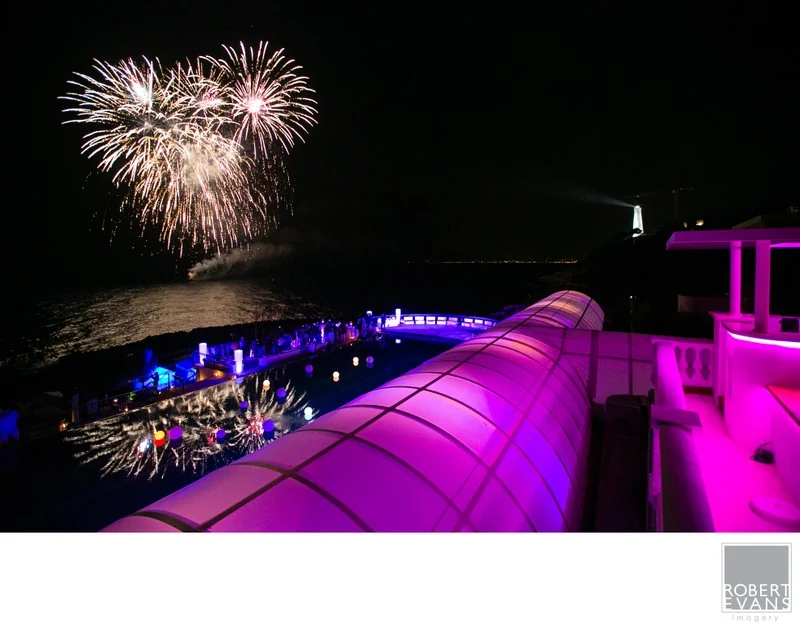 Fireworks above a nighttime celebration with vivid pink and purple architectural lighting.